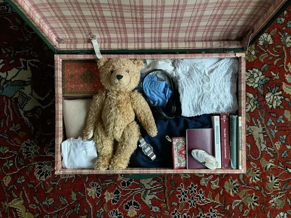 Открытый чемодан с вещами на ковре: плюшевый мишка, книги, часы